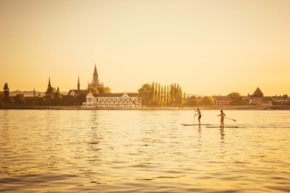Two persons stand-up paddleboarding on the calm waters of Lake Constance, surrounded by the warm glow of sunset