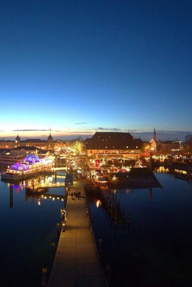 The brightly lit Christmas market in Konstanz at night, seen from the lake, with festive lights reflecting on the water